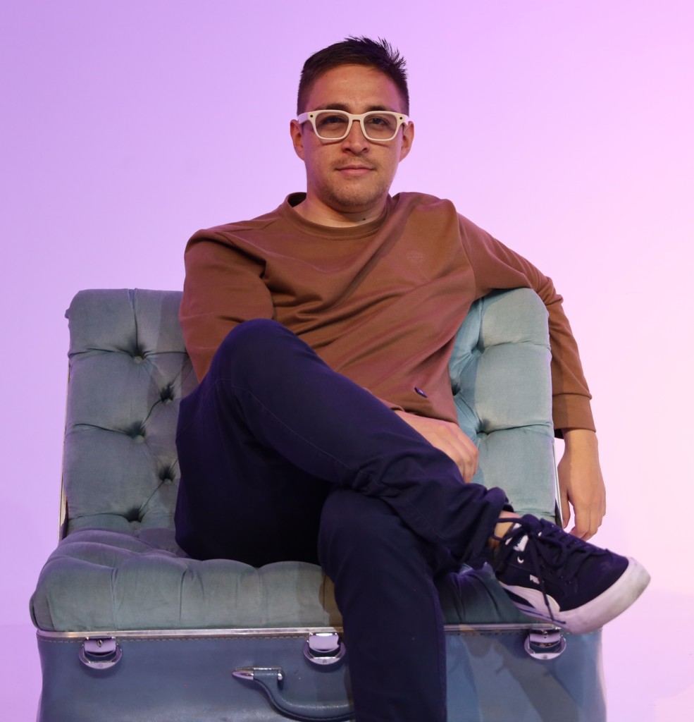 Camilo Guerrero con gafas blancas sentado en un sillón sobre fondo lila